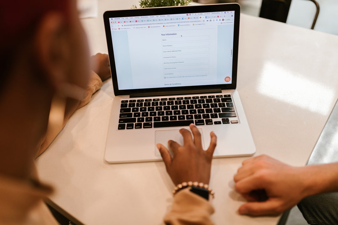 A person is using a laptop on a white table, filling out an online form. Another person’s hand is nearby, suggesting collaboration and learning.